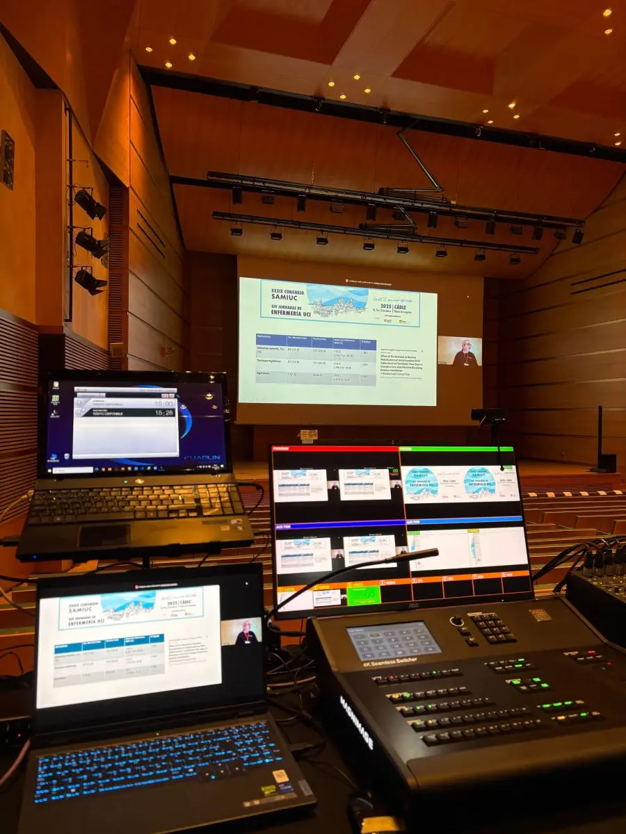 Puesto de control audiovisual con dos portátiles y una mesa de mezclas, con la sala y la pantalla al fondo