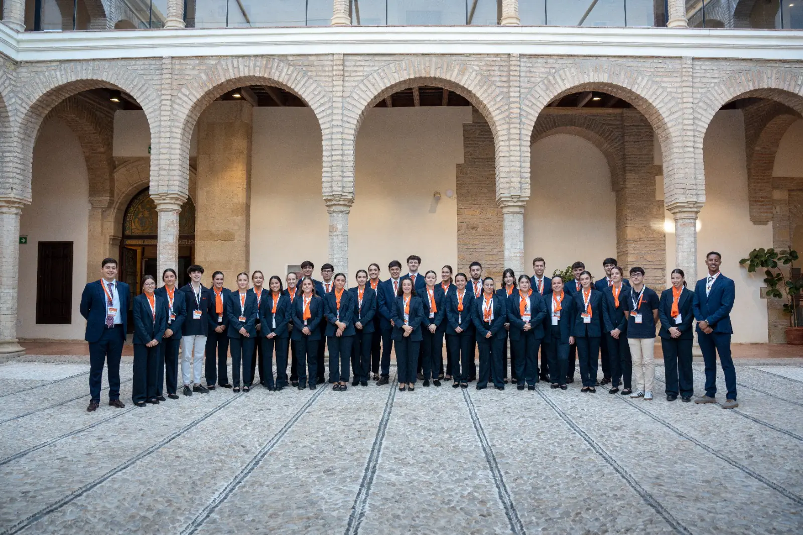 El equipo de azafatas y azafatos de Esmeeting, de traje azul marino y pañuelo naranja, posando en un patio con arcos de ladrillo